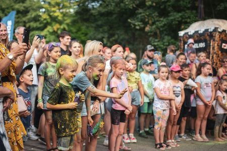 Grupa dzieci z bliska obserwuje występy, a mała dziewczynka w umazanej koszulce wyciąga rękę przed siebie