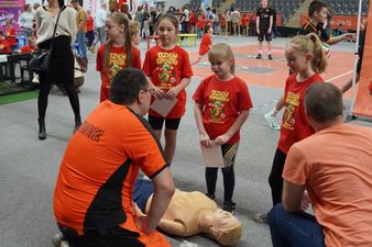 Cztery dziewczynki w koszulkach Misja Dinozaur stoją przed manekinem służącym do udzielania pierwszej pomocy podczas rozmowy z ratownikiem