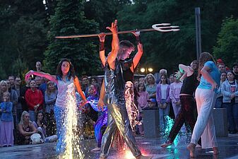 Artyści w błyszczących kostiumach tańczą wokół podświetlonej fontanny miejskiej podczas wieczornego pokazu. Mężczyzna w centrum trzyma trójząb.
