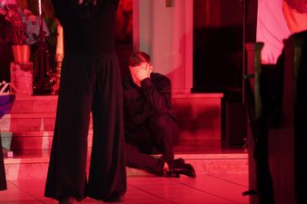 Mężczyzna ubrany na czarno siedzi na schodku, ręką zasłania twarz, z przodu osoba ubrana na czarno