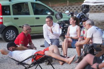 Grupa osób siedzi i rozmawia na ławeczkach na żwirowym podłożu. Mężczyzna w białej koszuli gestykuluje, a inny leży w leżaku obok nich. W tle widać jasnozielony samochód i motocykl