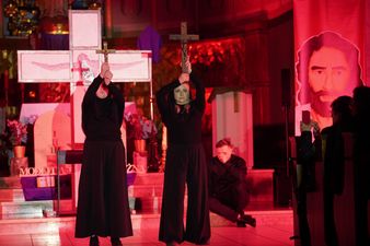 Aktorzy ubrani na czarno trzymają wysoko nad głowami krzyże, z boku obrazy Jezusa Chrystusa, w tle wizerunek krzyża