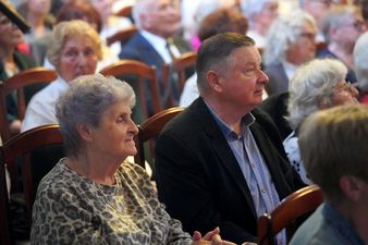 Starsza kobieta w bluzce i złotym łańcuszku obok mężczyzna w marynarce siedzą na krzesłach