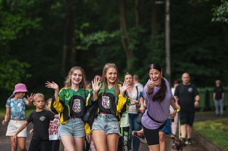 Maszerująca grupa uczestników imprezy plenerowej, na czele z uśmiechniętymi dziewczętami machającymi do aparatu