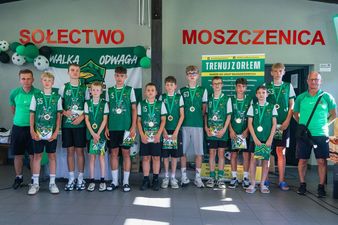 Grupa młodych piłkarzy w zielonych koszulkach z medalami na szyjach stoi w rzędzie z trenerami, a na ścianie za nimi widnieje napis „SOŁECTWO MOSZCZENICA” oraz baner z hasłem „TRENUJ Z ORŁEM”