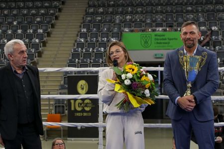 kobieta przemawia do mikrofonu w ringu bokserskim, trzymając bukiet kwiatów. Obok niej stoi mężczyzna w garniturze z dużym złotym pucharem