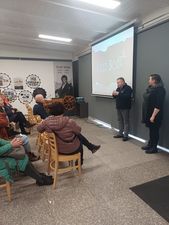 Ujęcie z boku uczestnicy spotkania siedzą na krzesłach na sali, patrzą na ekran