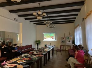 Stoły na który znajudją się prace plastyczne, obok uczestnicy warsztatów oglądający wyświetlony film.