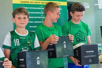 Trzech młodych piłkarzy w zielonych koszulkach stoi obok siebie, trzymając w dłoniach czarne pudełka z piłkami, podczas gdy jeden z nich przygląda się zawartości pudełka