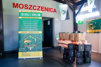 Stoisko promocyjne klubu UKS Orzeł Moszczenica z roll-upem zachęcającym do treningów, obok którego na stole znajdują się paczki dla zawodników oraz stos pudełek z odzieżą sportową