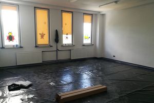 Wyremontowana sala z oknami zasłoniętymi żółtymi roletami z naklejkami. Podłoga zabezpieczona czarną folią, na której leżą materiały budowlane