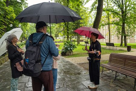 Spotkanie z przewodniczką w parku podczas deszczu. Uczestnicy wycieczki stoją na mokrej, kamiennej ścieżce i słuchają opowieści pod rozłożonymi parasolami – czarnym, białym oraz bordowym. W tle widać parkowe trawniki i drzewa