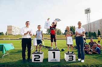Trzech chłopców stoi na podium zawodów rowerowych na stadionie, trzymając nagrody i dyplomy