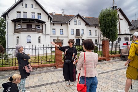 Kobieta pełniąca rolę przewodnika, ubrana w czarną narzutkę z motywem konia, wykonuje gest dłonią, opowiadając grupie o stojącym za ogrodzeniem dużym, białym budynku z kaplicą i krzyżem na dachu