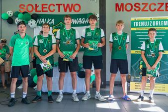 Pięciu młodych piłkarzy w zielonych strojach sportowych stoi w szeregu, każdy z nich trzyma w dłoniach małą torbę, a na szyi ma zawieszony medal
