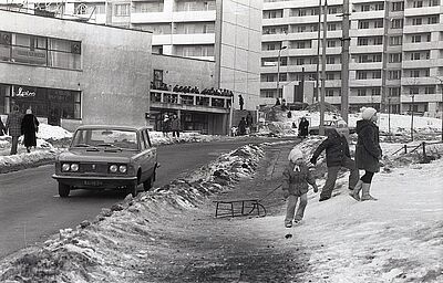 Zimowy widok osiedla miejskiego — dzieci bawiące się ze sankami na zaśnieżonym poboczu oraz jadący Fiat 125p