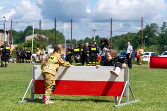 dziewczynka w piaskwym mundurze opiera się o biało-czerwoną przeszkode, dzieczywnka w czrnym mundurze próbuje ją pokonać