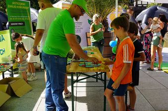 Stół integracyjny z grami edukacyjnymi, wokół grupka dzieci i dorosłych, z boku baner z napisem Jastrzębski Zakład Komunalny