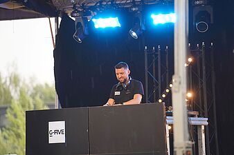Uśmiechnięty DJ występujący na scenie plenerowej za konsolą z logo „G-FIVE” w niebieskim oświetleniu