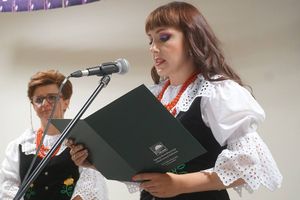 Dwie kobiety w strojach ludowych na scenie, jedna czyta podziękowania