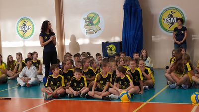 Uczniowie Zespołu Szkół Mistrzostwa Sportowego siedzą na parkiecie hali sportowej podczas uroczystości, w tle widoczne logo szkoły na ścianie