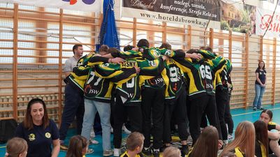 Grupa młodych sportowców w żółto-czarnych strojach z logo JSW stoi w kręgu, trzymając się za ramiona w geście drużynowej jedności na hali sportowej