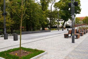 Szeroka, jasna promenada wyłożona kostką brukową. Po prawej stronie ciąg drewnianych ławek miejskich i wysokie, ciemne latarnie. Po lewej stronie młode drzewko w gruncie