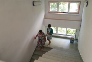 Dwie kobiety schodzą po nowo wykończonych, jasnych schodach na klatce schodowej. Ściany są białe, a pod oknem na półpiętrze stoi wiadro budowlane