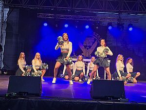 Zespół czirliderek w błyszczących bluzkach i ciemnych spódniczkach tańczący na oświetlonej scenie