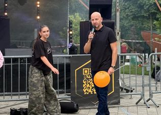 7. Mężczyzna stoi przed sceną z pomarańczowym balonem, mówiąc do mikrofonu. Po jego lewej idzie dziewczyna, patrząca wprost na aparat.