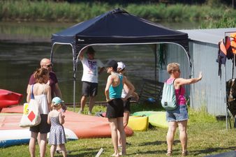 Grupa osób rozmawiająca na trawiastym brzegu rzeki w pobliżu czarnego namiotu ogrodowego i przygotowanych do zwodowania kajaków