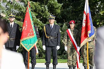 Uroczysty poczet sztandarowy, w skład którego wchodzą funkcjonariusze w ciemnych mundurach galowych (ze sztandarem) oraz kobieta w mundurze wojskowym trzymająca flagę Polski.