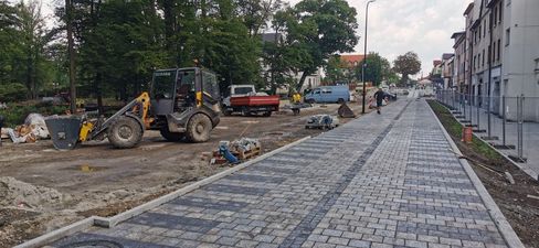 Nowo ułożona alejka z kostki brukowej w parku. Obok ładowarka kołowa i ciężarówka na placu budowy. W tle drzewa i ogrodzenie