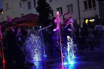 Wieczorny pokaz tańca z fontannami. Trójka artystów w ciemnych strojach porusza się wśród dynamicznych strumieni wody podświetlonych na niebiesko i różowo