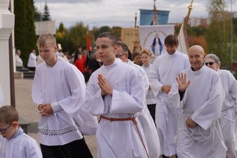 Duchowni w białych szatach biorą udział w procesji przed kościołem. W tle widać banery z wizerunkiem Matki Boskiej, flagi procesyjne oraz innych zgromadzonych biorących udział w procesji.