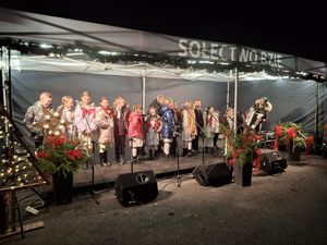 Grupa dzieci w strojach świątecznych występuje na scenie pod namiotem, obok nich stoi osoba grająca na akordeonie