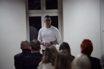 Młody mężczyzna w białej bluzie prowadzący prelekcję, z przodu grupa siedzących słuchaczy