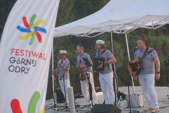 Biała flaga z kolorowym logo Festiwalu Górnej Odry po lewej stronie. Na dalszym planie zespół w pasiastych koszulkach gra koncert na pomoście przy wodzie