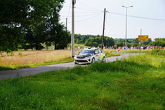 Samochód rajdowy jedzie wiejską, asfaltową drogą pośród pól i zieleni