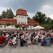 Ujęcie z boku, publiczność siedzi na krzesłach i słucha koncertu