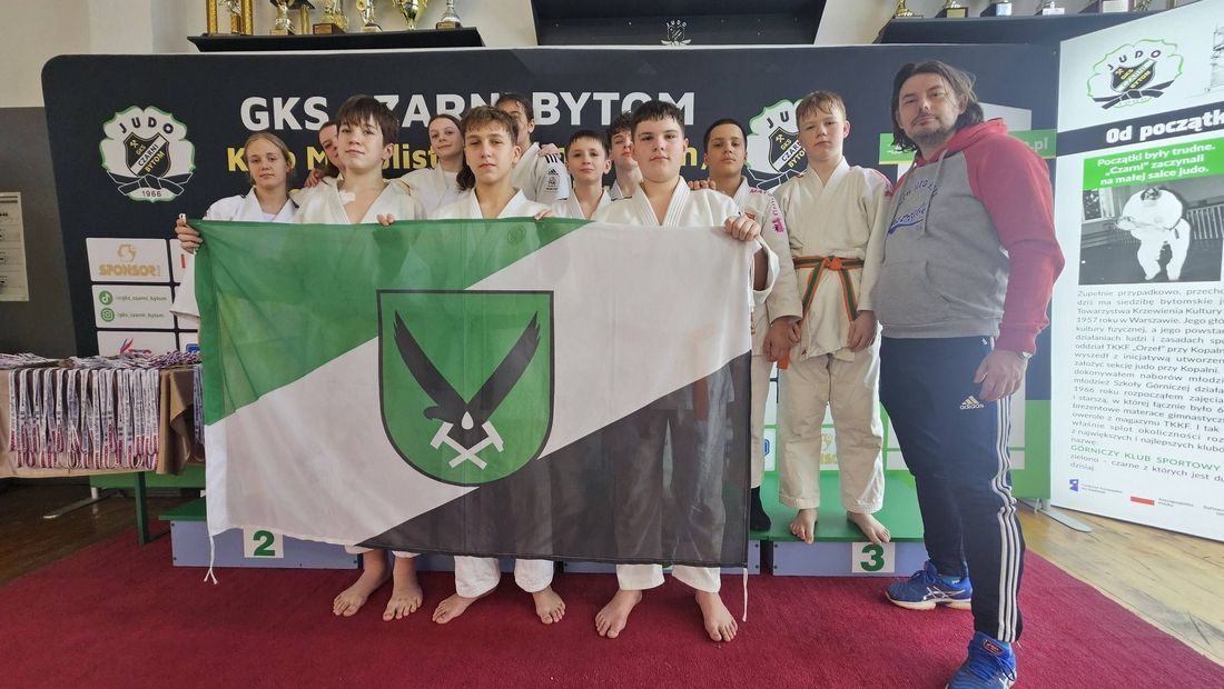 Grupowe zdjęcie drużyny Klub Judo Koka w strojach trzymający zieolono-biało-czarną flagę miasta. Obok stoi trener