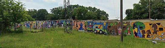 Długi murek pokryty wieloczęściowym muralem z postaciami z bajek (m.in. Stitch, pojazd ze Scooby-Doo), zrealizowanym na tle zielonej polany