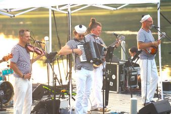 Czterej członkowie zespołu muzycznego w pasiastych koszulkach grają na instrumentach pod białym namiotem. Widoczny skrzypek, akordeonista, gitarzysta i mandolinista