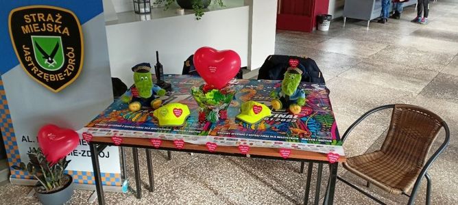 Stoisko Straży Miejskiej z Jastrzębia-Zdroju na finał WOŚP. Na kolorowym stole pluszowe krokodyle w mundurach, żółte czapki i czerwone serduszka