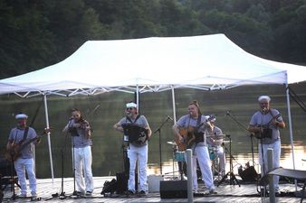 Występ zespołu szantowego na scenie przy jeziorze o zmierzchu. Muzycy grają na gitarach, skrzypcach, akordeonie i perkusji, ubrani w marynarskie pasiaki