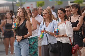 Uczestnicy plenerowego wydarzenia z drinkami w dłoniach obserwujący występ na scenie
