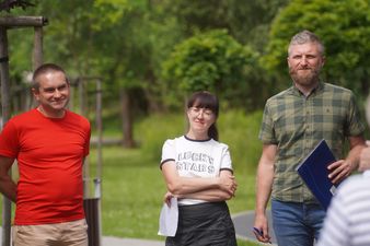 Trzy osoby stoją na tle zieleni: mężczyzna w czerwonym t-shircie, kobieta w białej koszulce ze skrzyżowanymi ramionami i brodaty mężczyzna w koszuli w kratę, który trzyma folder. Wszyscy są przedstawicielami organizatorów