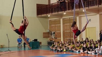 Zawodniczki w czerwonych strojach prezentują figury gimnastyczne na szarfach podczas występu przed publicznością szkolną