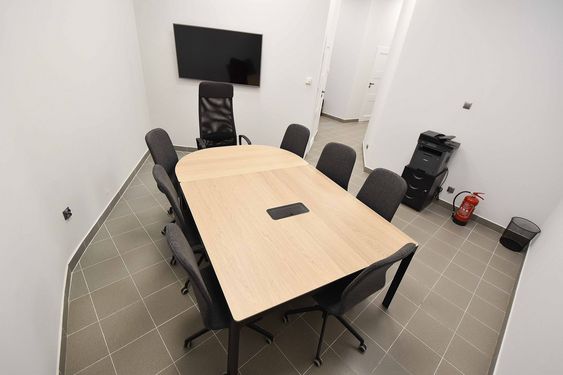 Sala konferencyjna z jasnym, drewnianym stołem i siedmioma ciemnymi krzesłami. Na białej ścianie wisi duży monitor. W rogu widoczna drukarka, gaśnica i kosz. Nowoczesne, sterylne wnętrze