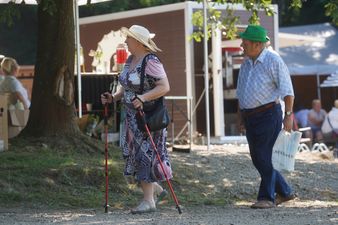 Starsza kobieta w letnim kapeluszu idąca z kijkami do nordic walking oraz towarzyszący jej mężczyzna w zielonym kapeluszu na plenerowym wydarzeniu